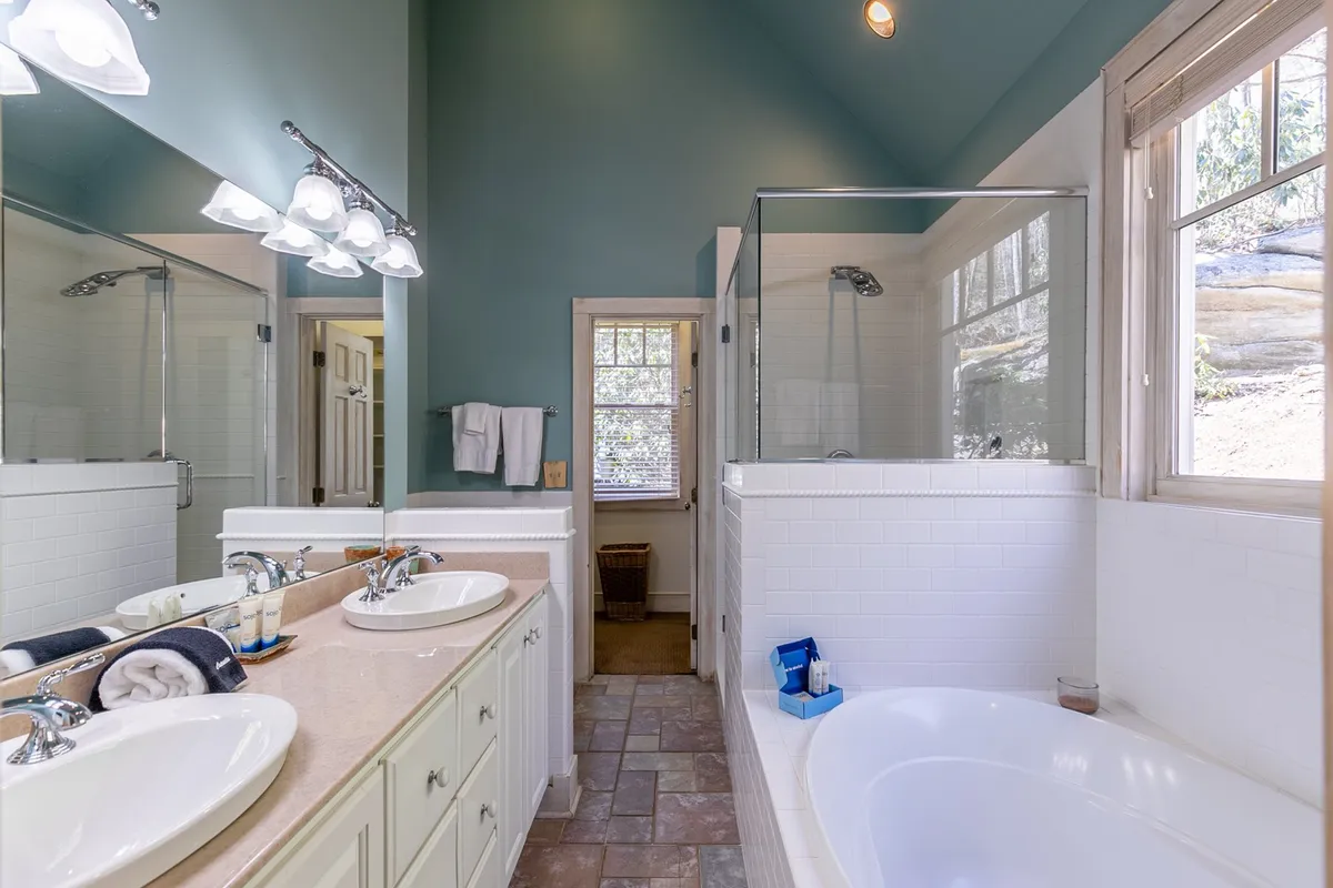 A luxurious bathroom with double vanity sinks, a glass shower enclosure, a freestanding soaking bathtub, and large windows that bathe the space in natural light. The setting is spa-like and tranquil, ideal for relaxation and morning routines.