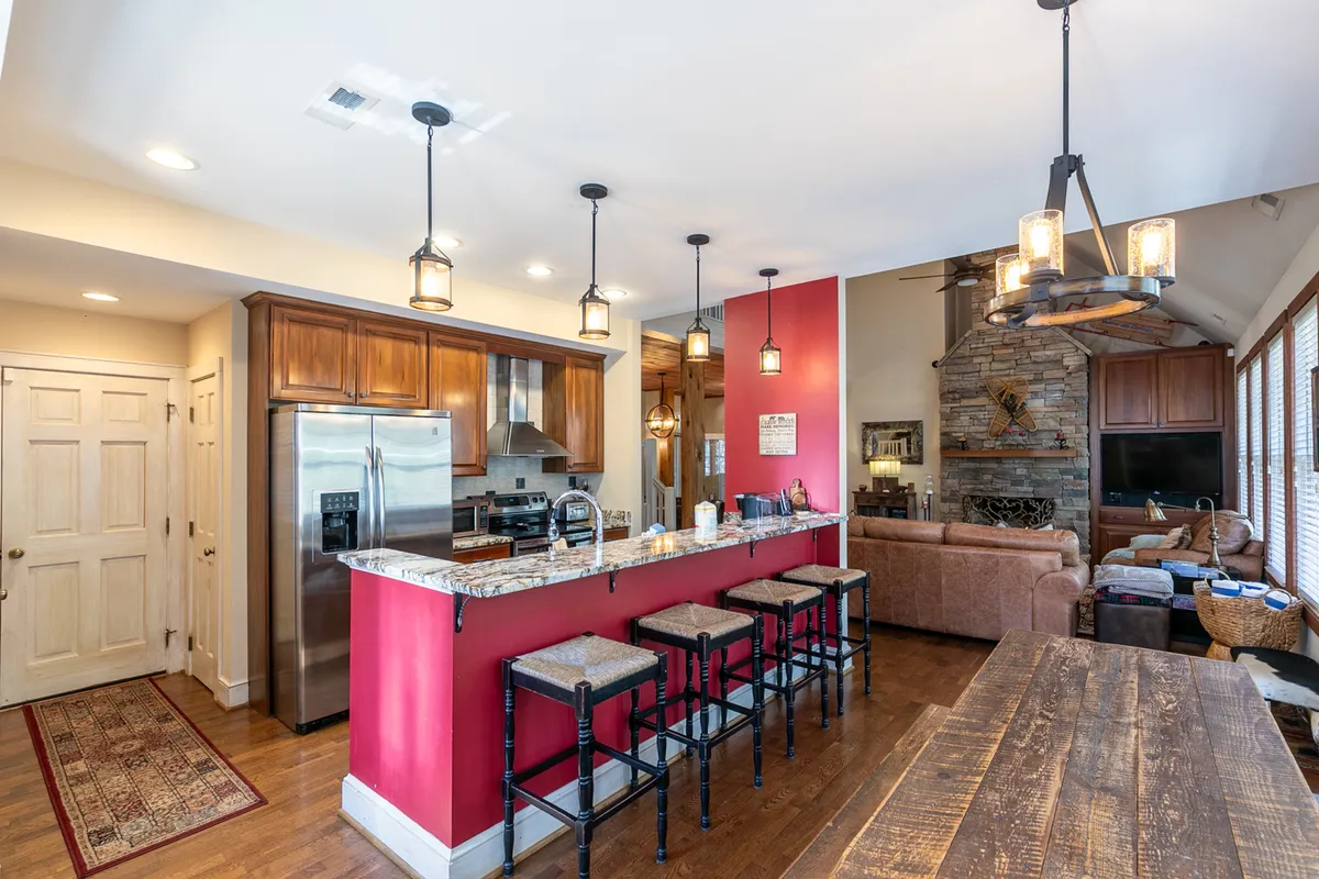 This image highlights the open-concept kitchen and dining area, featuring granite countertops, stainless steel appliances, a wooden dining table, red accent walls, and a view of the adjacent stone fireplace. Bar stools and a spacious layout emphasize the kitchen's role as a social hub for cooking and entertaining.