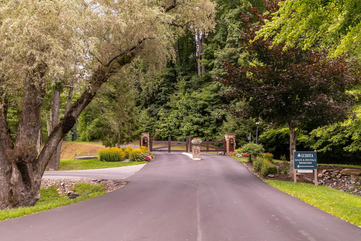 This image captures the gated entrance to the property, framed by floral landscaping, tall trees, and an inviting asphalt driveway. The scene sets a tranquil, welcoming tone, with natural greenery and flowers suggesting both privacy and an upscale, well-maintained environment.
