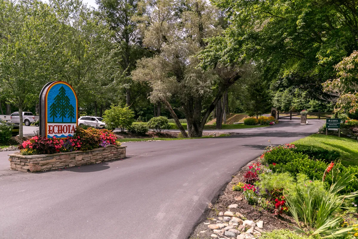 A welcoming entrance to the Echota community, marked by a prominent sign, landscaped flower beds, and a line of trees. The scene conveys a sense of arrival in a well-kept, nature-focused environment.