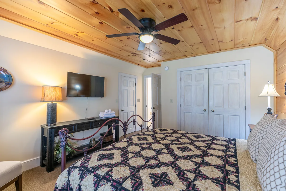 A bedroom with a quilted bedspread, wooden ceiling, ceiling fan, and a TV. The cozy setting offers a peaceful space for rest and includes entertainment options for extended stays.