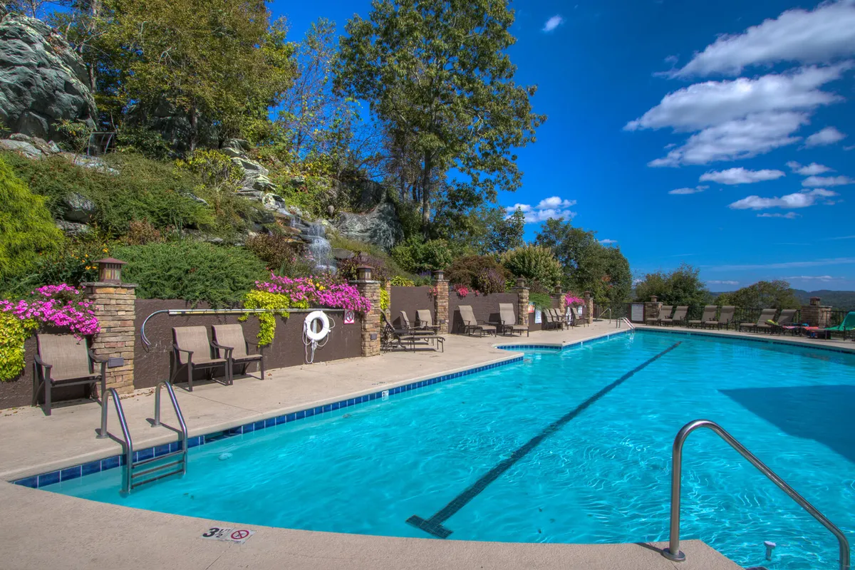 A sunlit outdoor swimming pool lined with lounge chairs and backed by a landscaped garden and a waterfall feature. The pool area is inviting and clearly family-oriented, suggesting both relaxation and fun activities for all ages.