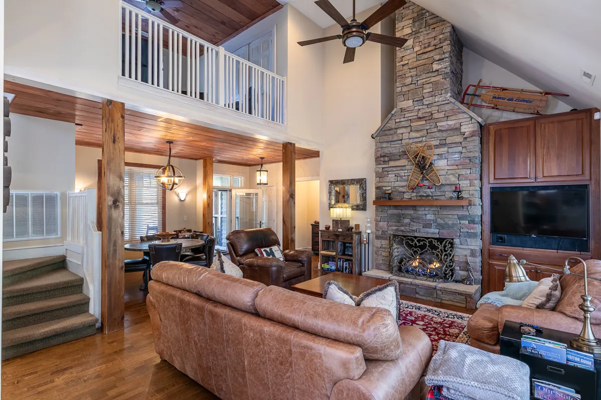 This inviting interior image captures the great room's soaring vaulted ceilings, rich wood floors, a stunning stone fireplace, plush leather sofas, and a dining area in the background. The wooden beams and warm lighting evoke a cozy, welcoming atmosphere perfect for gatherings and relaxation.
