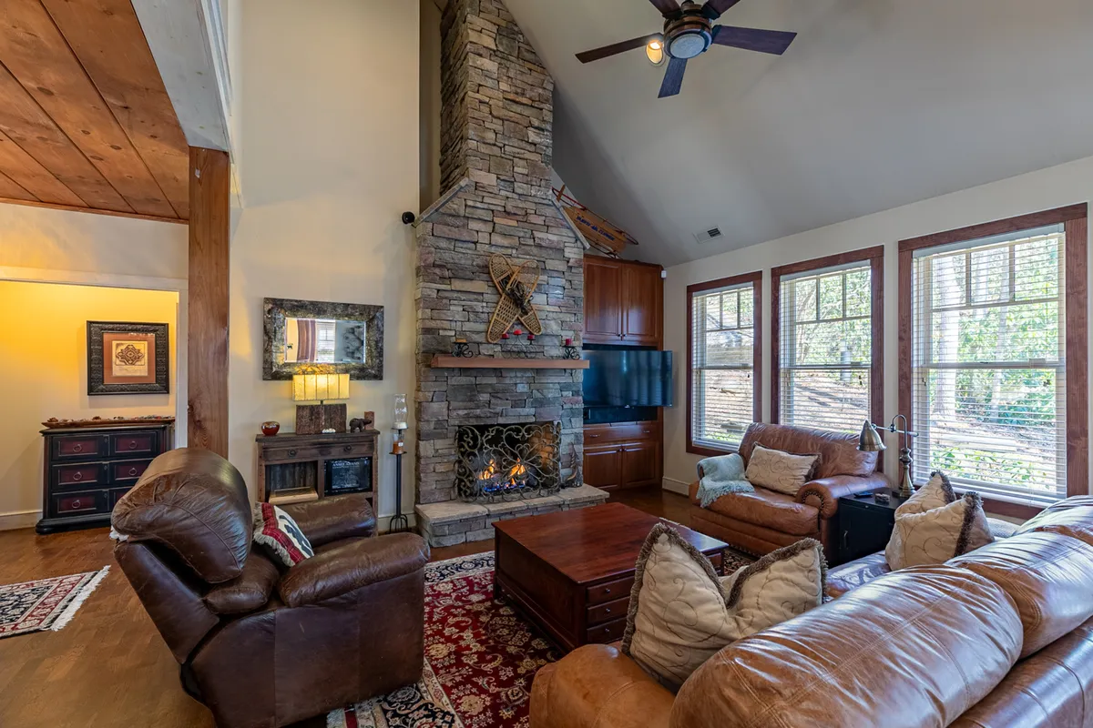 The image showcases a spacious great room with soaring wooden vaulted ceilings, a striking stone fireplace, plush leather sofas, and large windows that flood the space with natural light, creating a cozy and inviting atmosphere for relaxation and gatherings.