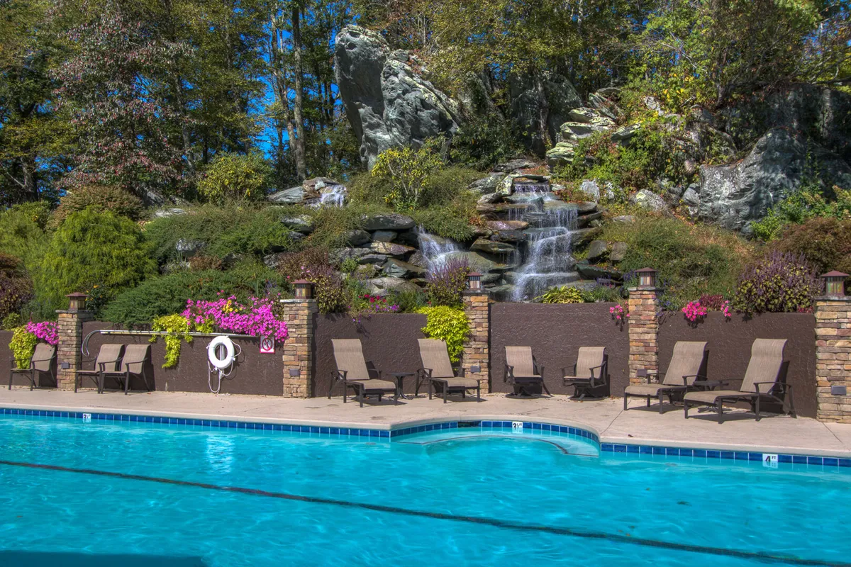 A tranquil outdoor swimming pool scene featuring a sparkling pool with a cascading waterfall, surrounded by lush landscaping, lounge chairs, and colorful flowers. The setting is perfect for sunbathing or relaxing by the water, with a rejuvenating, upscale resort ambiance.
