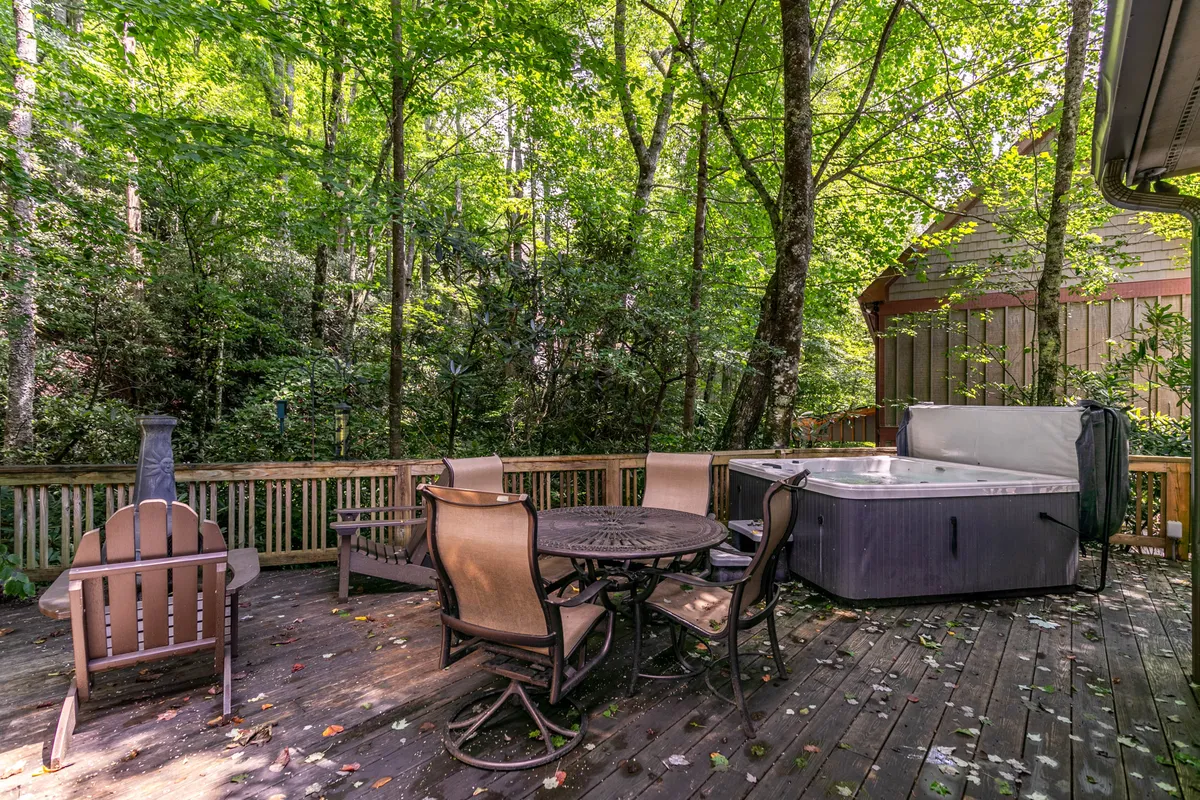 A peaceful wooden deck nestled among tall trees, featuring a bubbling hot tub, a cozy outdoor seating area, and natural surroundings. The hot tub is a focal point, inviting relaxation, while the deck offers space for mingling and enjoying the fresh mountain air.