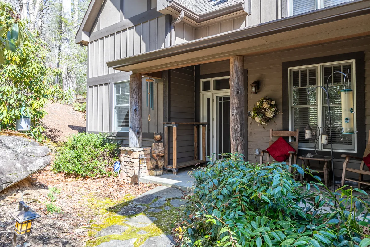 An inviting exterior scene shows the stone pathway leading to the home's entrance, flanked by a charming wooden bear statue and cozy outdoor seating. The decorative wreath on the door and natural elements create an atmosphere of warmth and hospitality.
