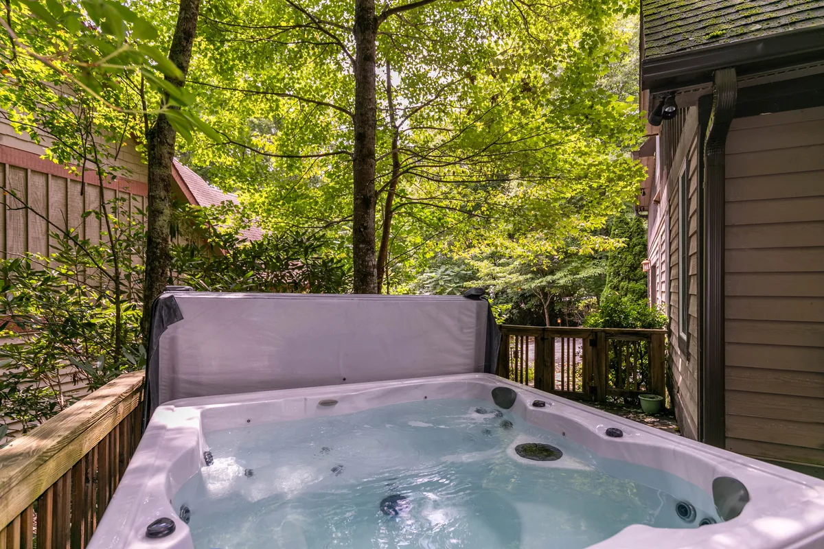 A private wooden deck nestled in greenery, featuring a bubbling hot tub and comfortable seating. The space is tranquil and perfect for unwinding outdoors after a day of activities.
