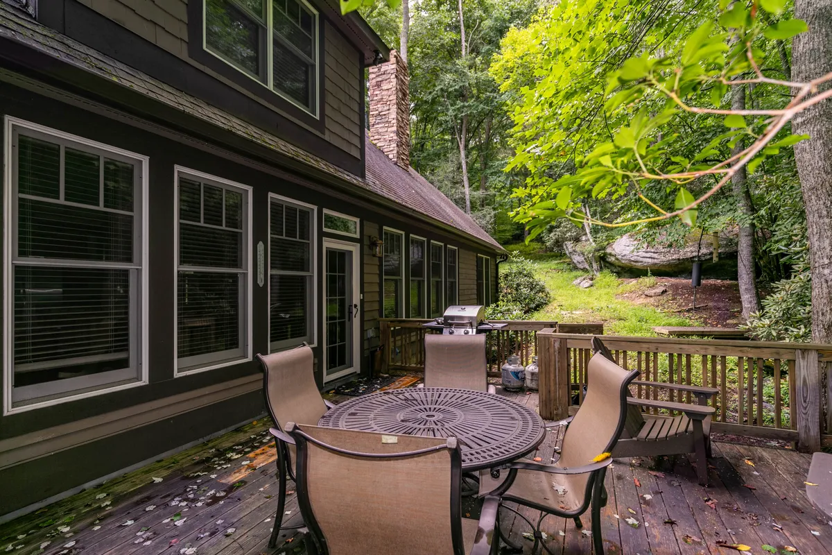 An inviting outdoor dining area set on a wooden deck, surrounded by mature trees. A large table is ready for alfresco meals, and a gas grill stands nearby, hinting at the promise of lively barbecues and sunset dinners surrounded by nature.